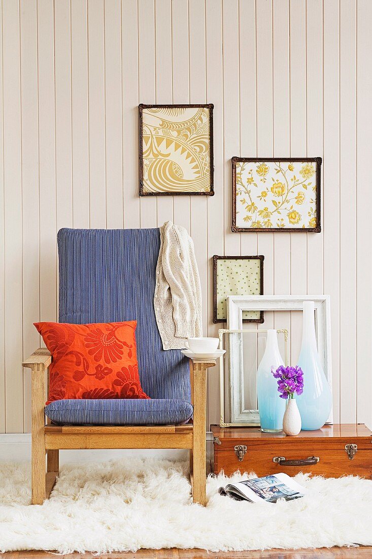 Orange patterned cushion on blue armchair with wooden frame, vases and empty picture frame on suitcase on floor below framed pictures on white wooden wall
