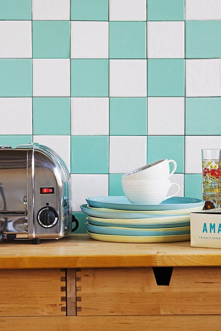 Stack of blue and yellow plates next to stainless steel toaster on wooden cabinet against turquoise and white chequered wall tiles