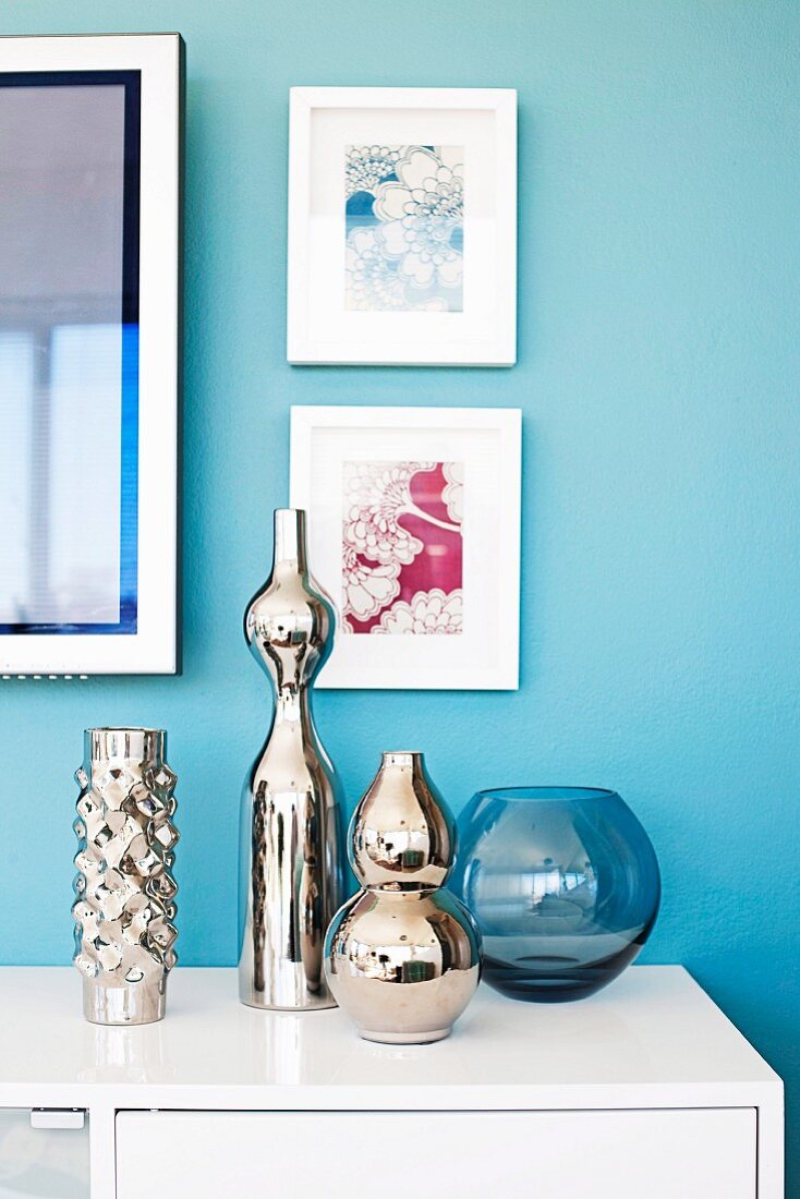 Verspiegelte Vasen in verschiedenen Grössen auf weißem Sideboard vor hellblau getönter Wand mit gerahmten Bildern