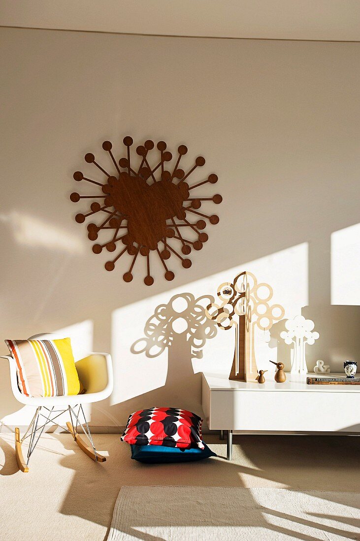 Artistic wall decoration and tree sculptures on a white lowboard next to floor cushions and a rocking chair