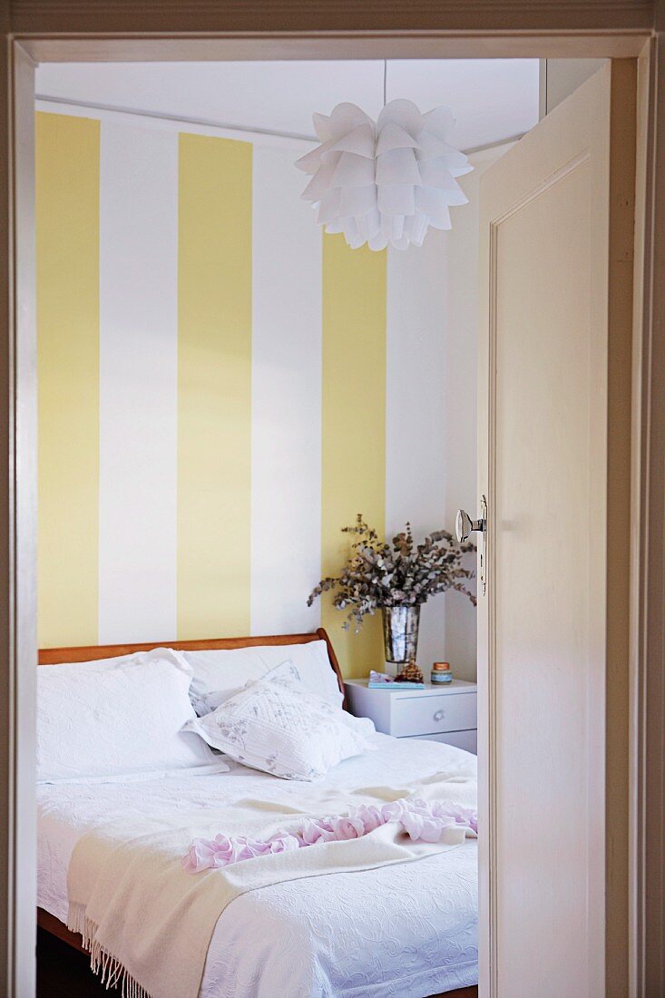 Blick durch offene Tür ins Schlafzimmer auf Bett mit weisser Bettwäsche, unter Bauhaus Hängeleuchte, an Wand gelbweiss gestreifte Tapete
