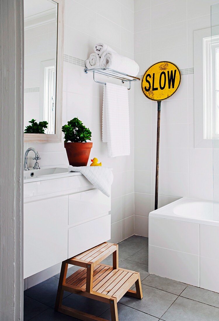 White wall tiles, wooden step stool, grey tiled floor, vintage traffic sign and towels on shelf in modern bathroom