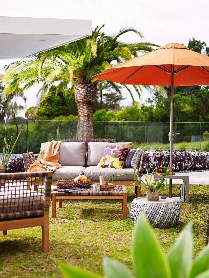Terrassenplatz vor Palme und Glasbrüstung, orangefarbener Sonnenschirm, Sofa und Geflechtmmöbel mit Retroflair