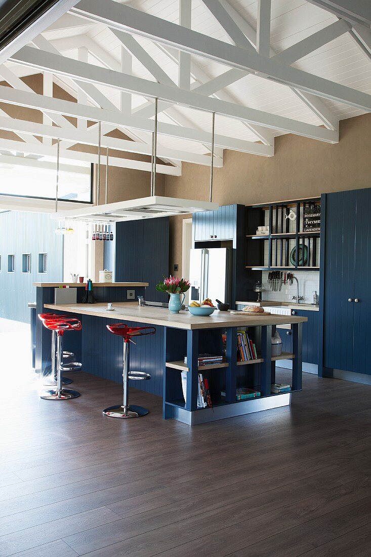 Kitchen counter with bar stools on wooden floor in high-ceilinged, open-plan interior with white-painted roof structure
