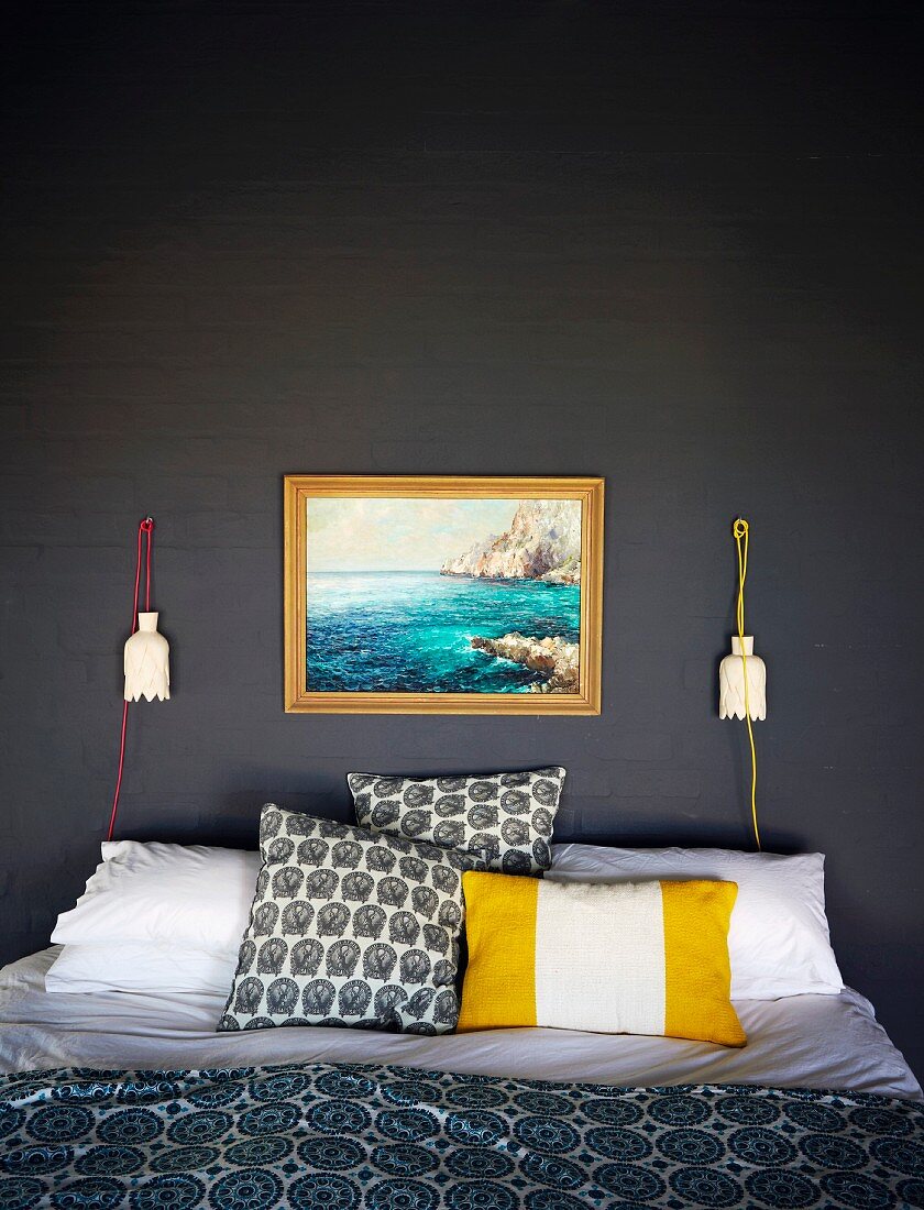 Verschiedene Kissen auf Doppelbett vor dunkelgrau gestrichener Ziegelwand mit Landschaftsbild und künstlerischen Wandleuchten