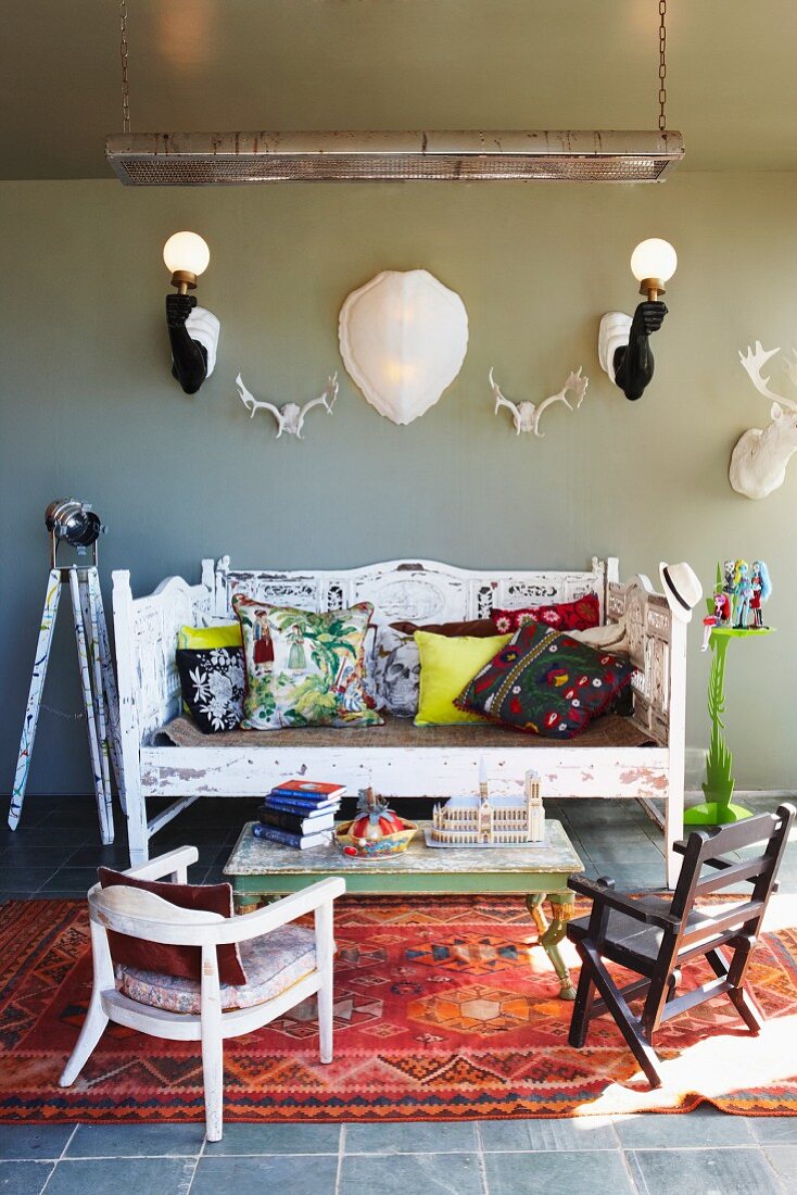 Verschiedene Stühle und Couchtisch auf folkloristischem Teppich vor Sitzbank an grauer Wand mit Wandleuchte zwischen Tiertrophäen