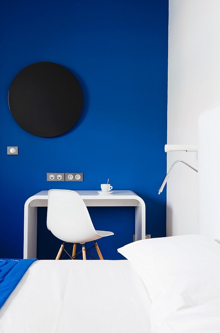 Farbstyling im Schlafzimmer mit Designer Schreibplatz und Klassikerstuhl vor königsblau getönter Wand mit schwarzer Scheibe