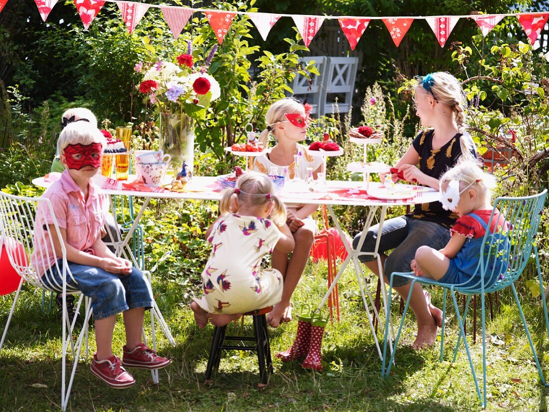 Kinder mit Masken sitzen an gedeckten Partytisch im Garten