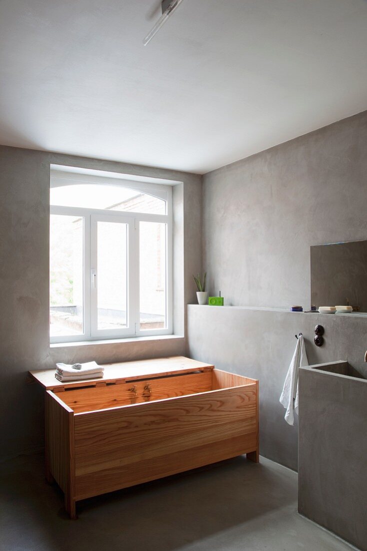 Designerbadewanne aus Holz mit offenem Deckel vor restauriertem Badfenster, seitliche Wand mit Vorsatzschale und Waschtisch in Betonausführung