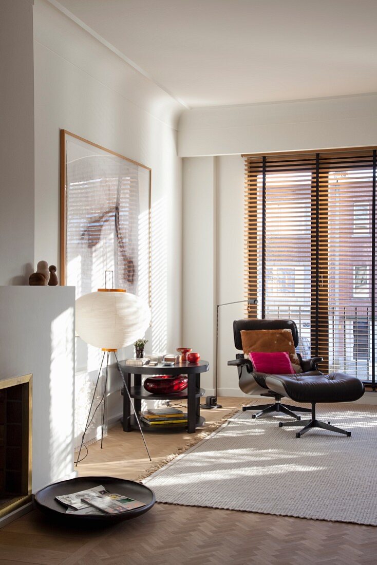 Eames Lounge Chair and matching footstool on pale rug in corner of living room in front of balcony doors with louver blinds; wooden dish on floor in front of open fireplace in foreground