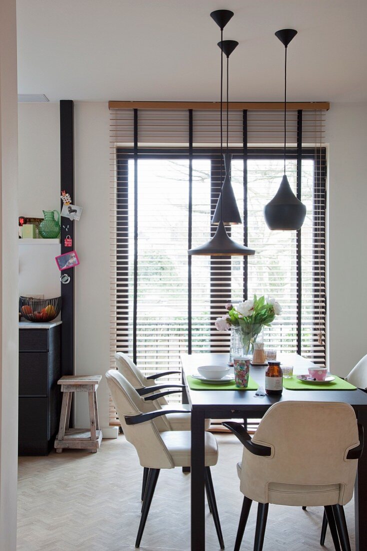 Chairs covered in white leather at black dining table set for breakfast below designer pendant lamps; balcony doors with louver blinds in background