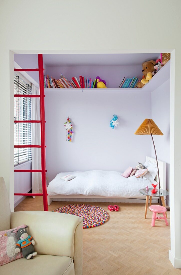 Partially visible armchair in front of red ladder, standard lamp with pleated lampshade next to bed and books and soft toys on high encircling shelf
