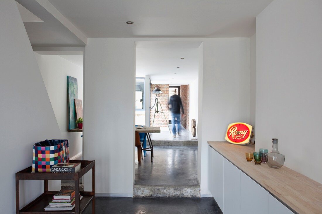 Offene minimalistische Küche mit Blick durch raumhohen Durchgang in nachfolgende Räume mit unterschiedlich hohen Podesten und einer Person im Hintergrund