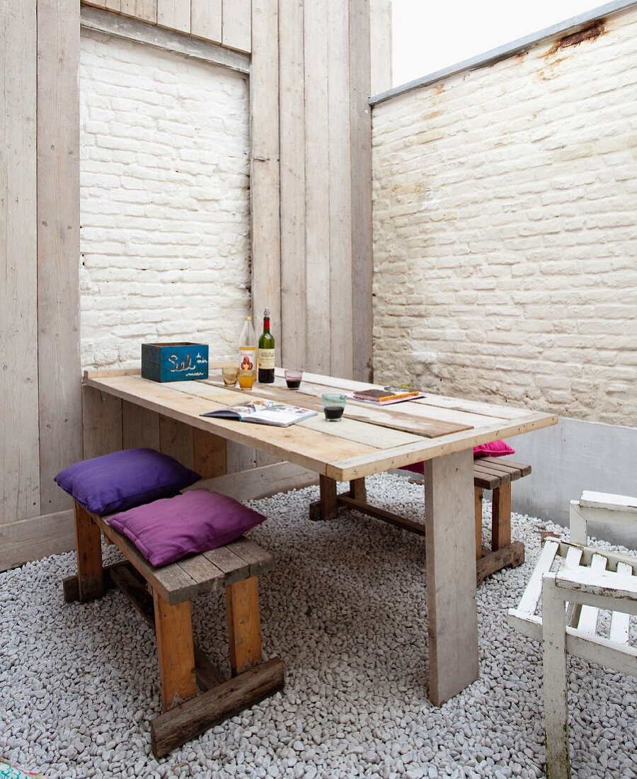 Fold-up wooden table and benches with colourful cushions on patio with whitewashed brick walls and gravel floor