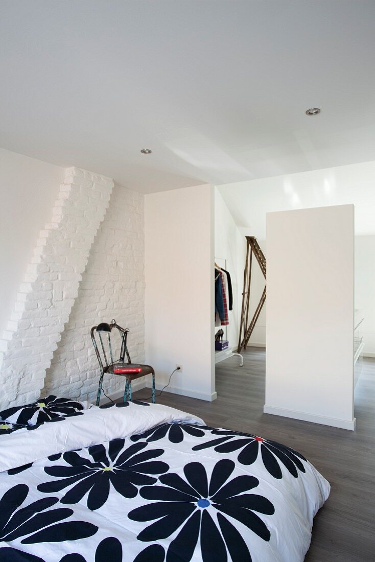 Geblümte Bettwäsche auf schlichtem Bett im Dachzimmer, weiße Raumteiler vor offener Ankleide mit minimalistischem Flair