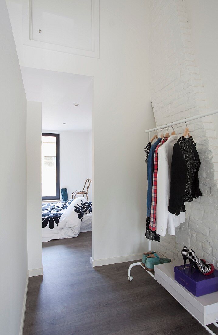 Clothes rack with shoe rack on castors against whitewashed brick wall and view of bed and window through floor-to-ceiling doorway
