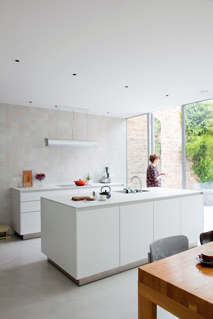Minimalist, white, designer kitchen with tiled wall and open glass wall leading to terrace with tall brick garden wall