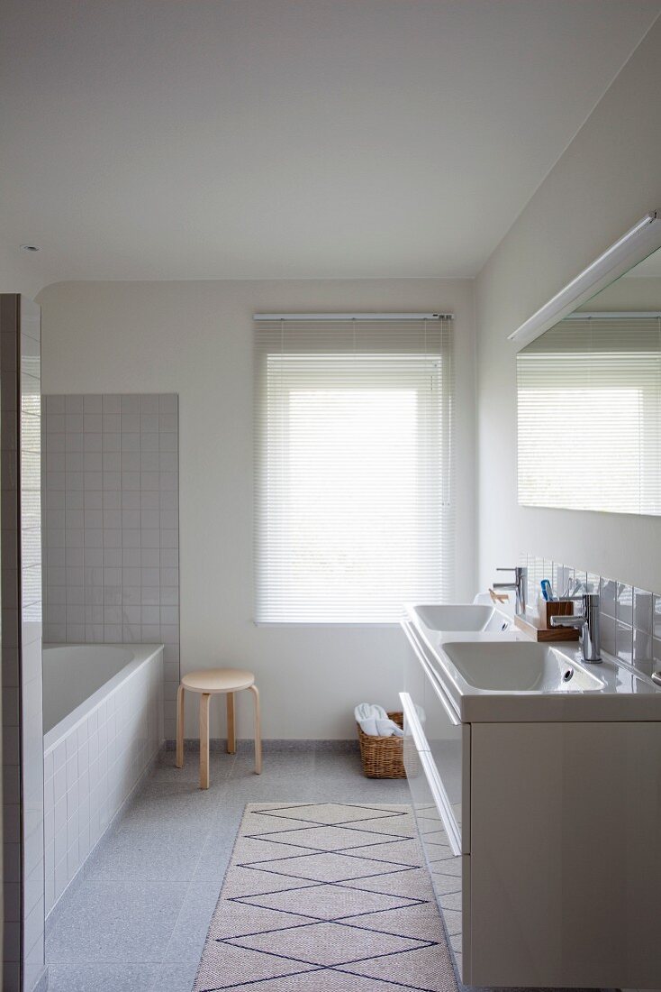 Minimalist bathroom with pale grey tiles, white bathtub, twin sinks, window with blind and rug next to washstand base unit