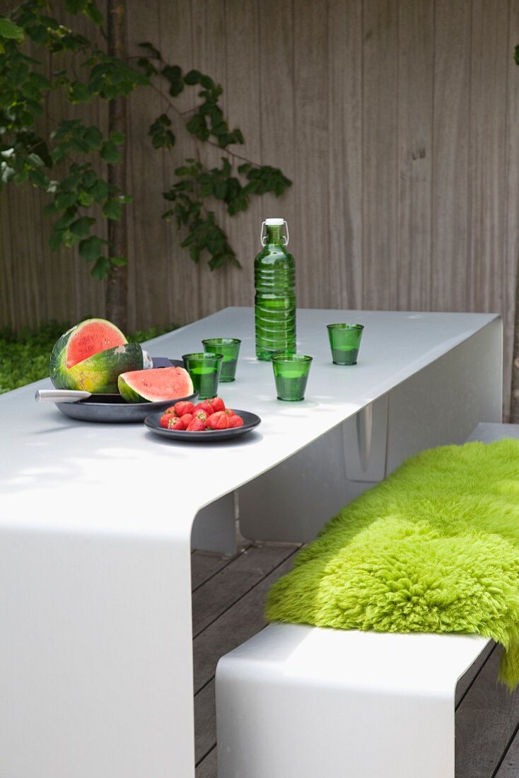 Frisches Obst und Getränk in grüner Flasche mit passenden Gläsern auf weißem Tisch, davor Sitzbank mit grüner Auflage auf der Terrasse