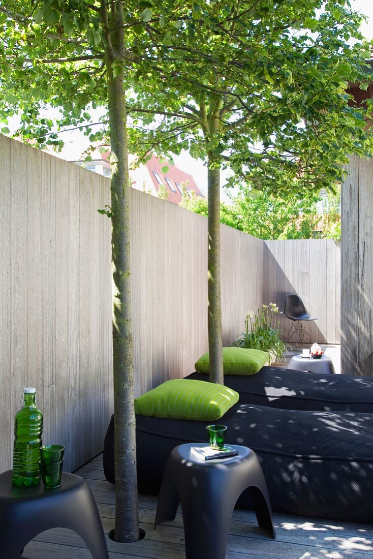 Schwarze Polsterliegen mit grünem Kissen und Kunststoff-Hocker auf der Terrasse vor Holzzaun