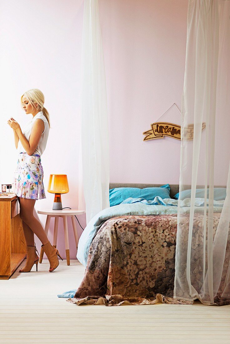 Blonde woman standing at console table next to double bed with airy curtains attached to suspended rods