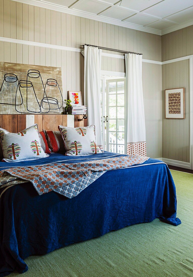 Double bed with blue bedspread and scatter cushions against wood-clad wall and green rug in bedroom
