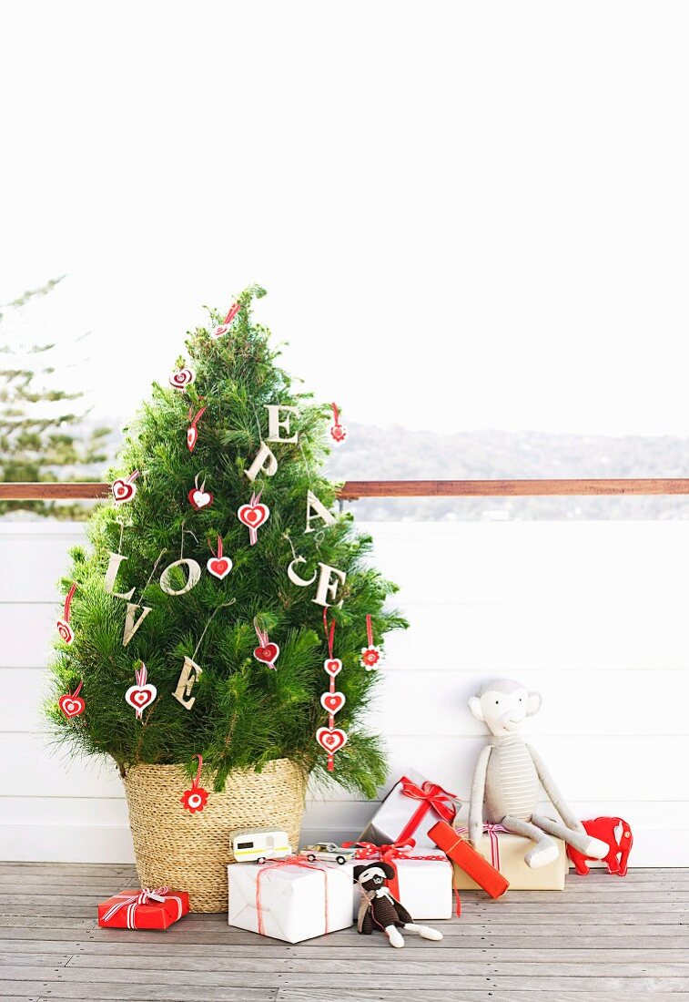 Kieferbäumchen mit Holzbuchstaben und herzförmigen Anhängern im Strohkorb, daneben Weihnachtsgeschenke und Spielzeuge