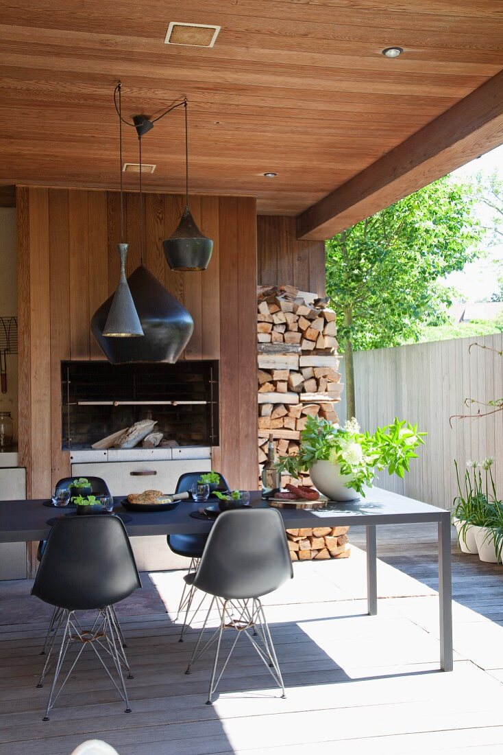 Schwarze Schalenstühle mit filigranem Metallgestell vor langem Tisch auf überdachter Terrasse gegenüber Kamin und Holzlager an Holzwand im Innenhof