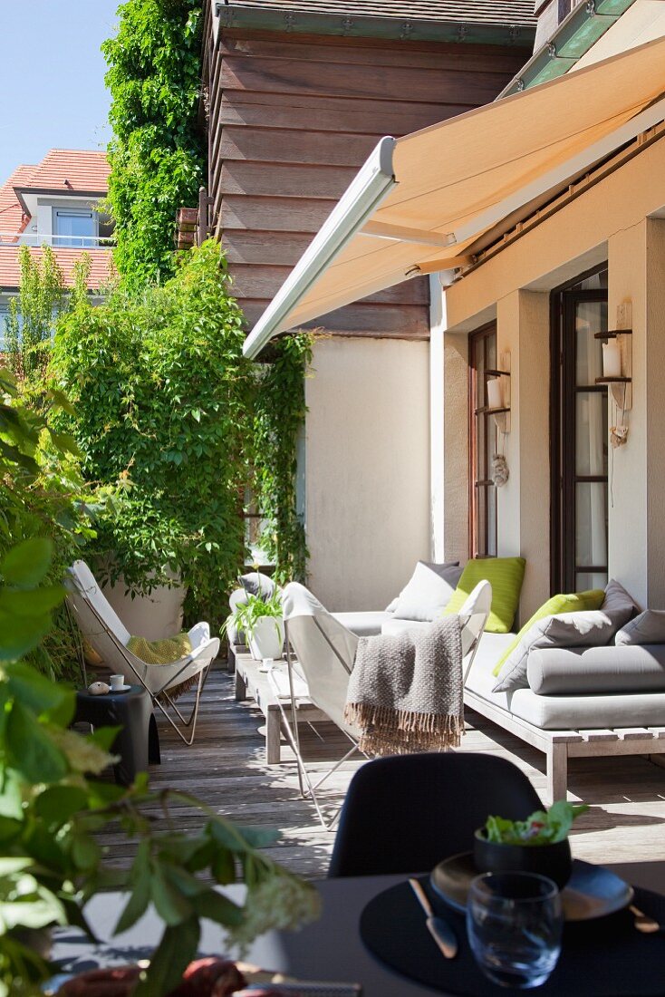 weiße Butterfly Chairs um Couchtisch und schlichte Holzbank mit Polstern auf sonnenbeschienener Terrasse vor Wohnhaus mit Markise