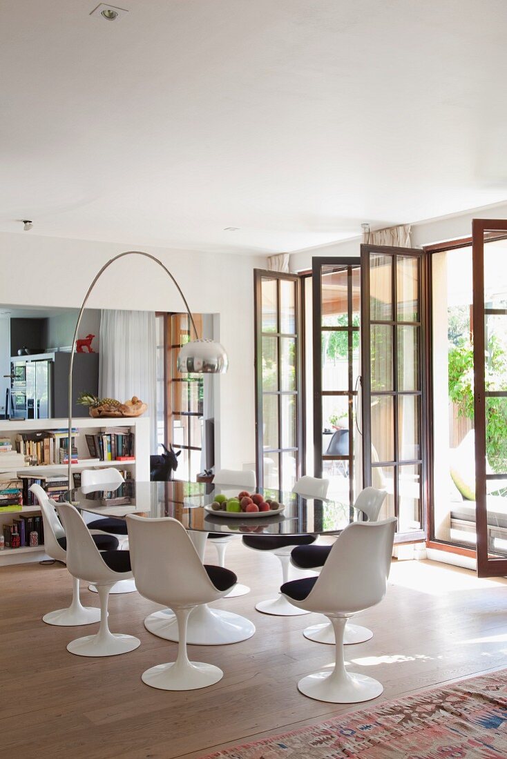 White tulip chairs with dark seat cushions around oval glass table below arc lamp and open lattice French windows to one side in modern interior