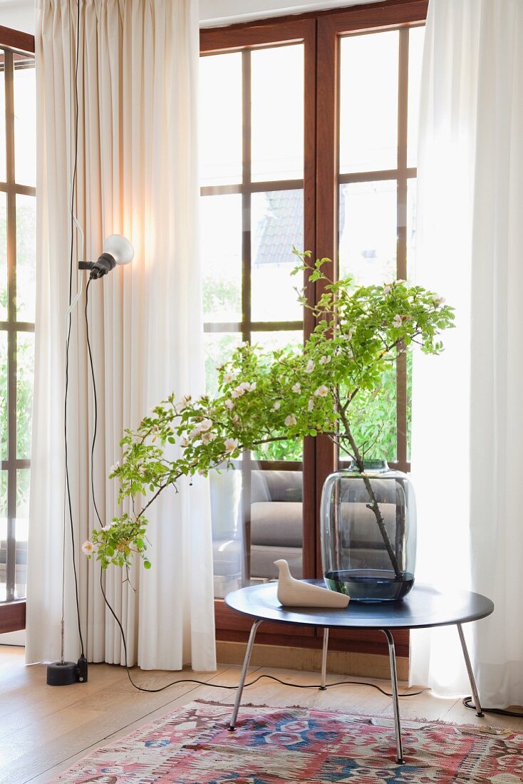 Branch of leaves in glass vase on round side tale and standard lamp in front of lattice French windows with airy curtains
