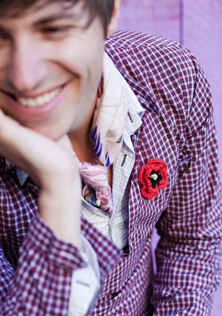 Lachender junger Mann mit roter gehäkelter Schmuckblume als Anstecker an lila kariertem Hemd