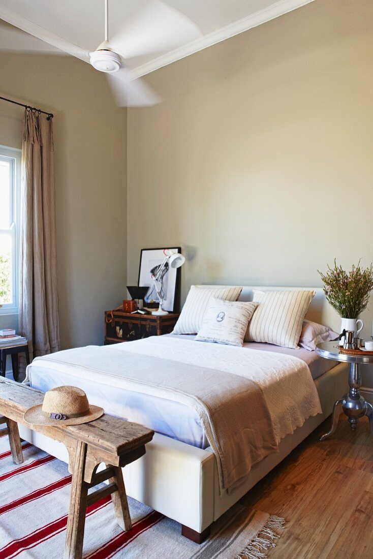 Doppelbett mit Plaid und Kissenstapel vor grau getönter Wand, an Bettende rustikale Holzbank