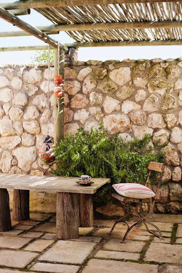 Vintage- Gartenstuhl mit Kissen und rustikale Holzbank auf Steinboden einer Terrasse mit Bambus Pergola vor hoher Natursteinmauer