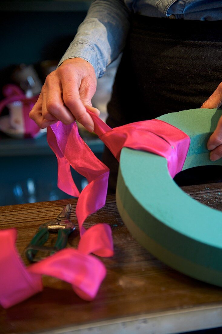 Frauenhände beim Gestalten eines Blumenkranzes mit Steckschaum und pinkfarbenem Seidenband