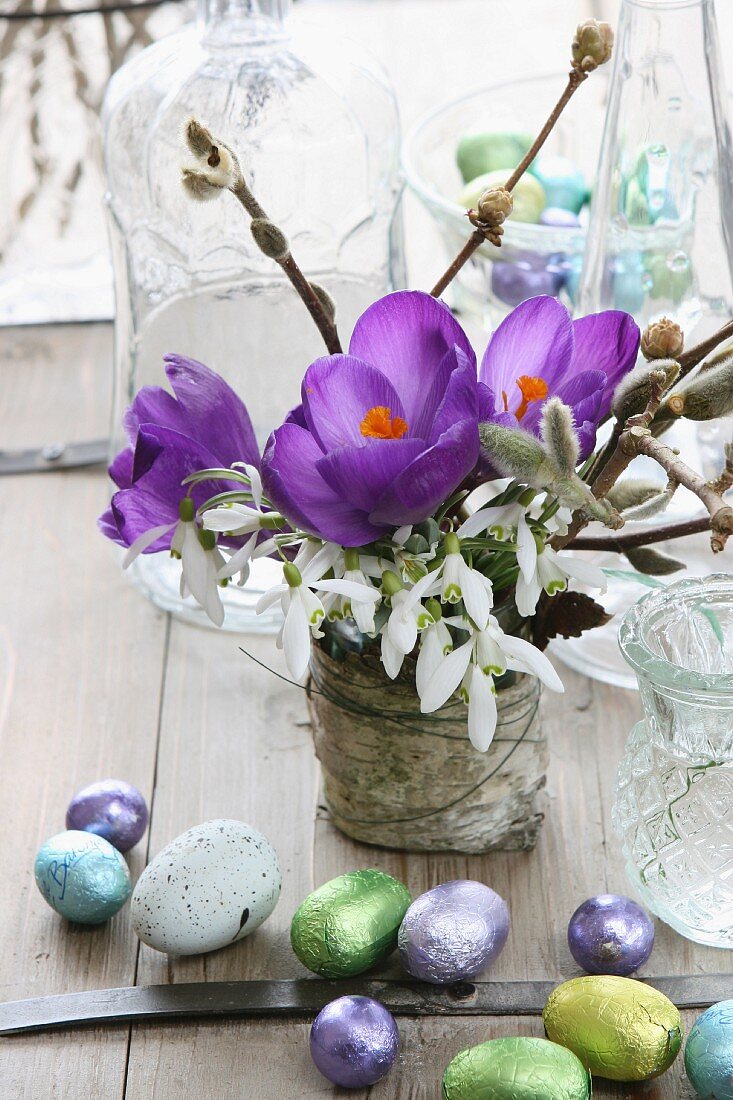 Frühlingsstrauss mit lila Krokussen, Schneeglöckchen und Weidenkätzchen, daneben Schokoladenostereiern