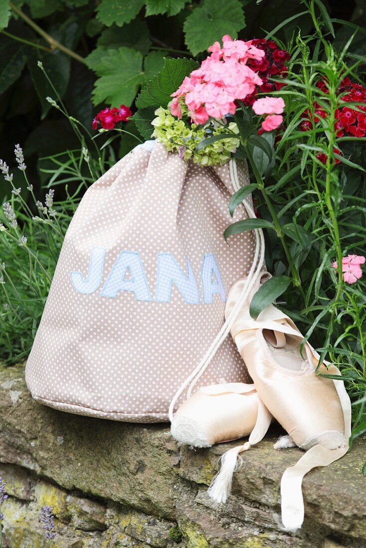 Hand-sewn, polka dot sports bag with sewn-on name next to ballet shoes on stone wall in garden