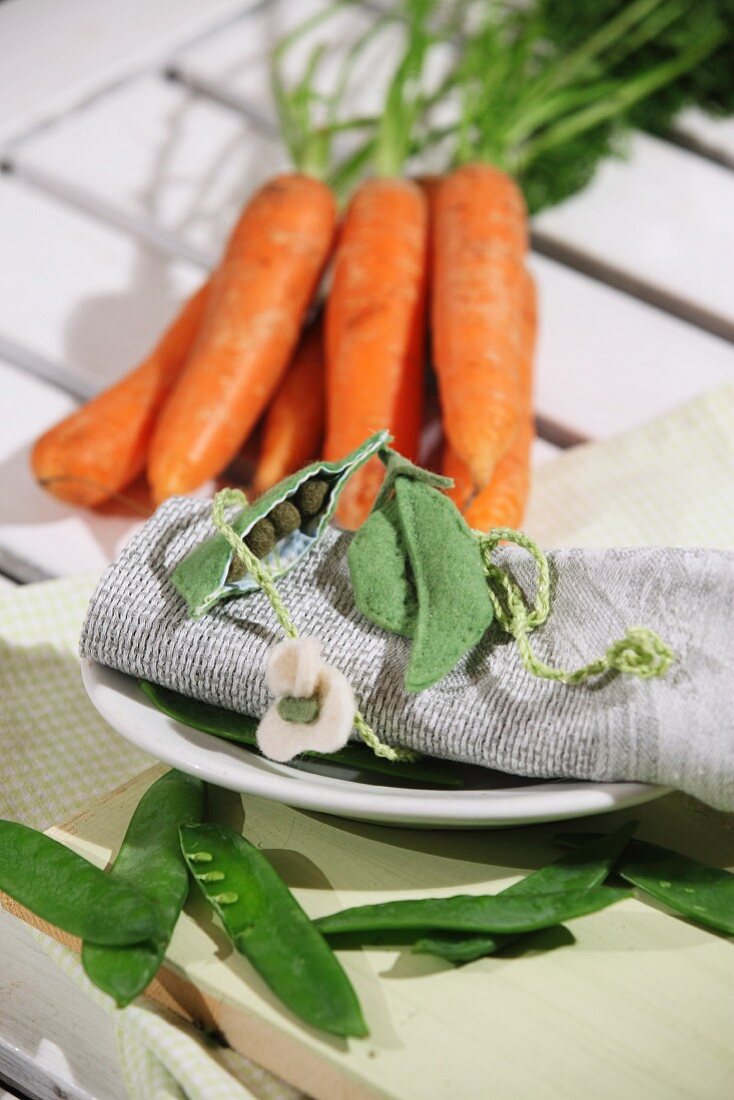 Felt pea pods arranged on rolled linen napkin in front of bunch of carrots
