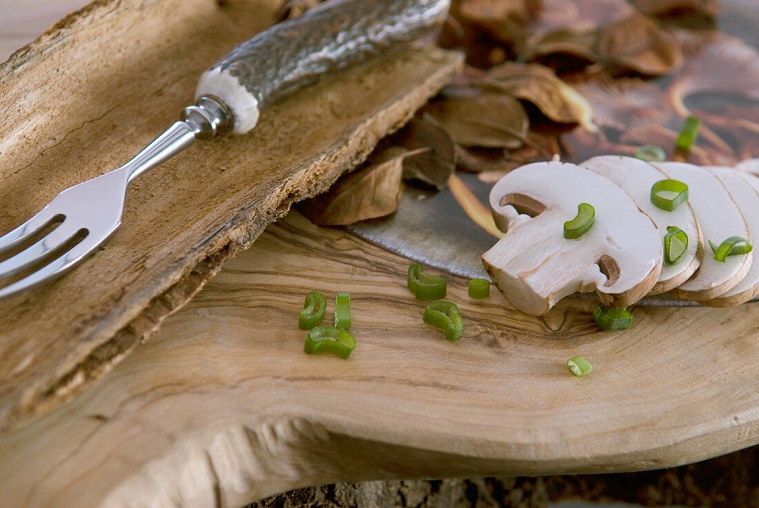 Aufgeschnittene Champignons und Frühlingszwiebel auf Holzbrett; rustikale Gabel mit Horngriff in einer Rindenschale