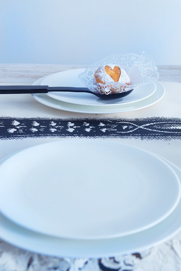 weiße Teller und schwarzer Löffel mit Gebäck auf Spitzenpapier und bedruckter Tischläufer aus weißem Papier