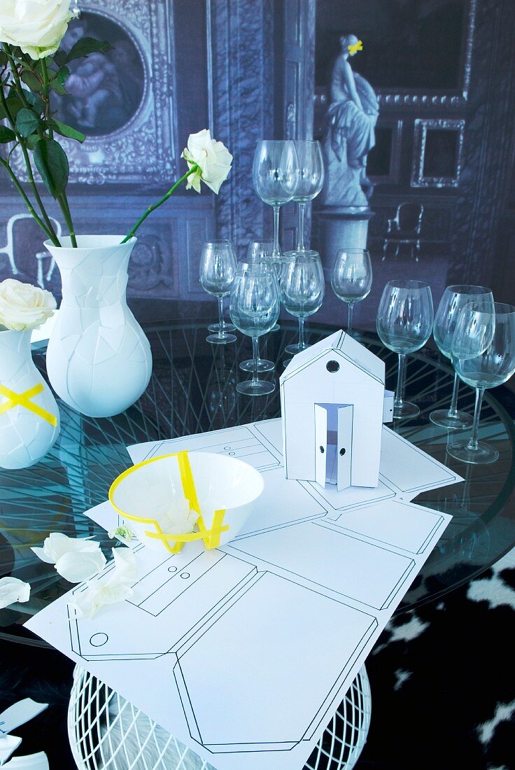 Symbolen für eine Hochzeit: Haus zum Selberplanen aus Karton und zerbrochenes Geschirr mit gelbem Klebeband beklebt, im Hintergrund Stielgläser und Blumenvasen auf Glastisch