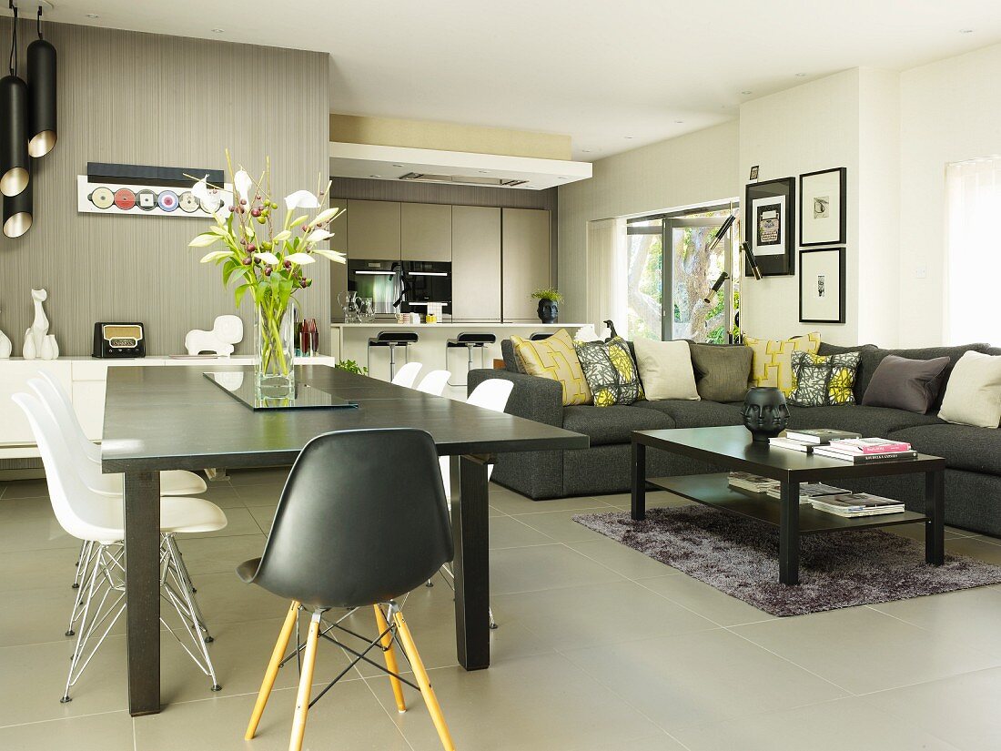 Schalenstühle und grauer Esstisch neben Loungeecke mit Überecksofa, dahinter Kochbereich in modernem, offenem Ambiente mit grauem Fliesenboden