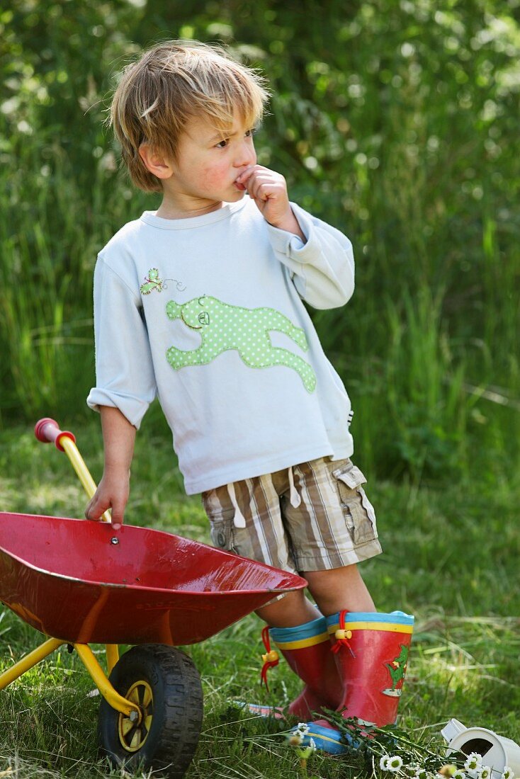 Kind mit selbstgenähtem Froschmotiv auf Shirt neben roter Schubkarre auf der Wiese