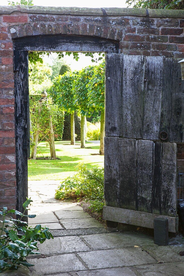 Offene Holztür in Ziegelmauer und Blick auf Baumreihe in englischem Garten