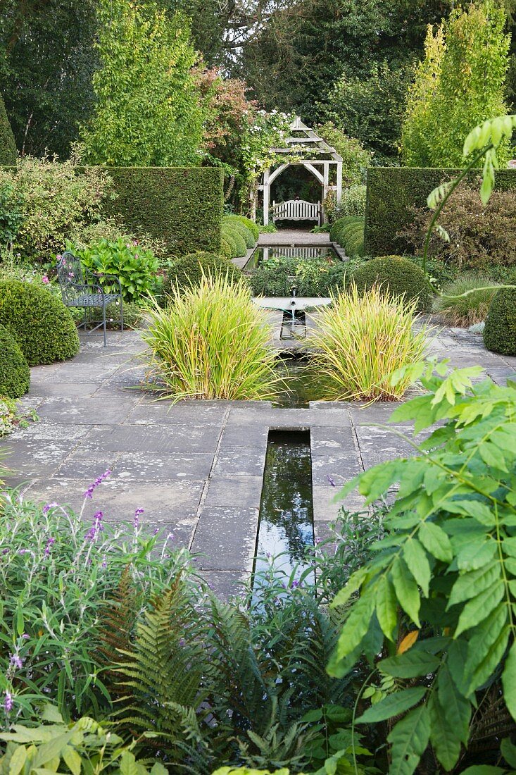 Steinboden mit schmalen, eingelassenen Wasserrinnen und Ziergräser, im Hintergrund Wasserbecken vor traditionellem Pavillon in englischem Garten