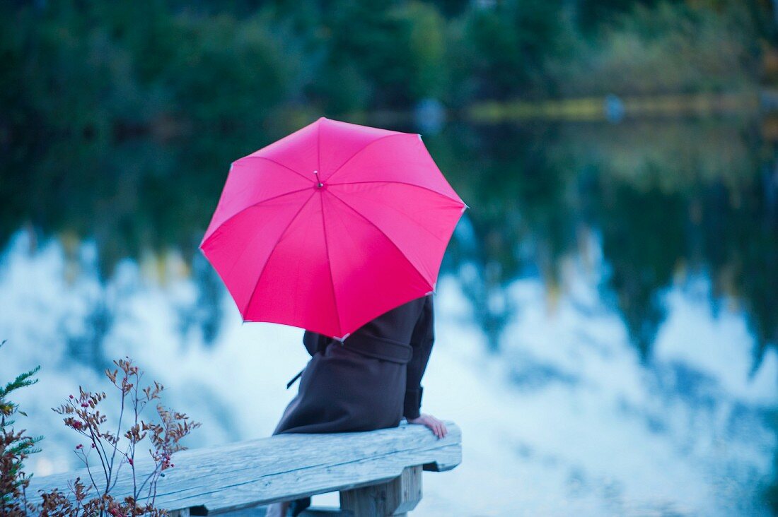 Frau mit pinkfarbenem Regenschirm am See sitzend