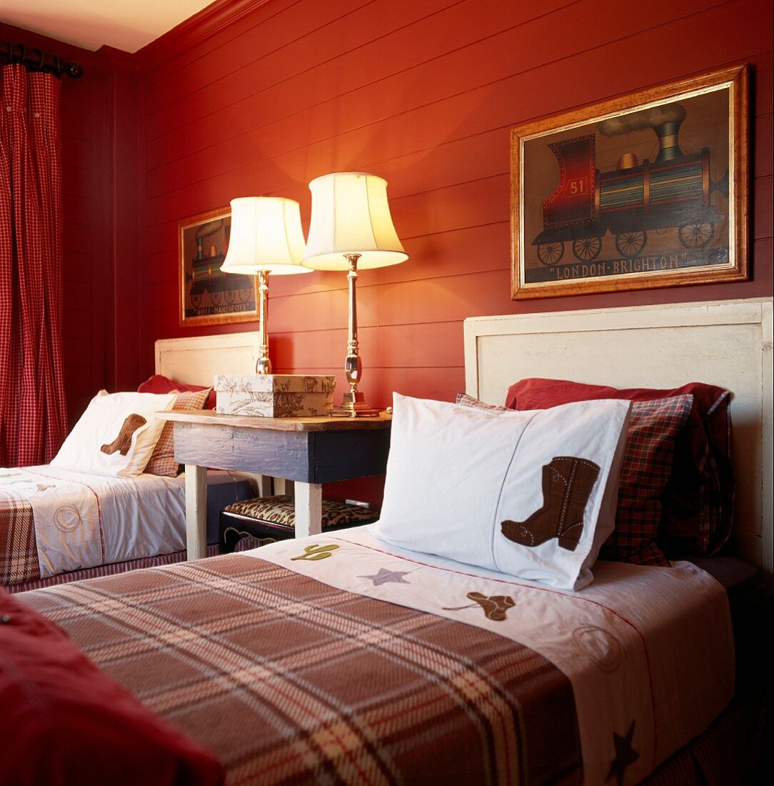 Twin country-house beds with tartan blankets against red-painted wooden wall and traditional bedside lamps with brass bases in bedroom
