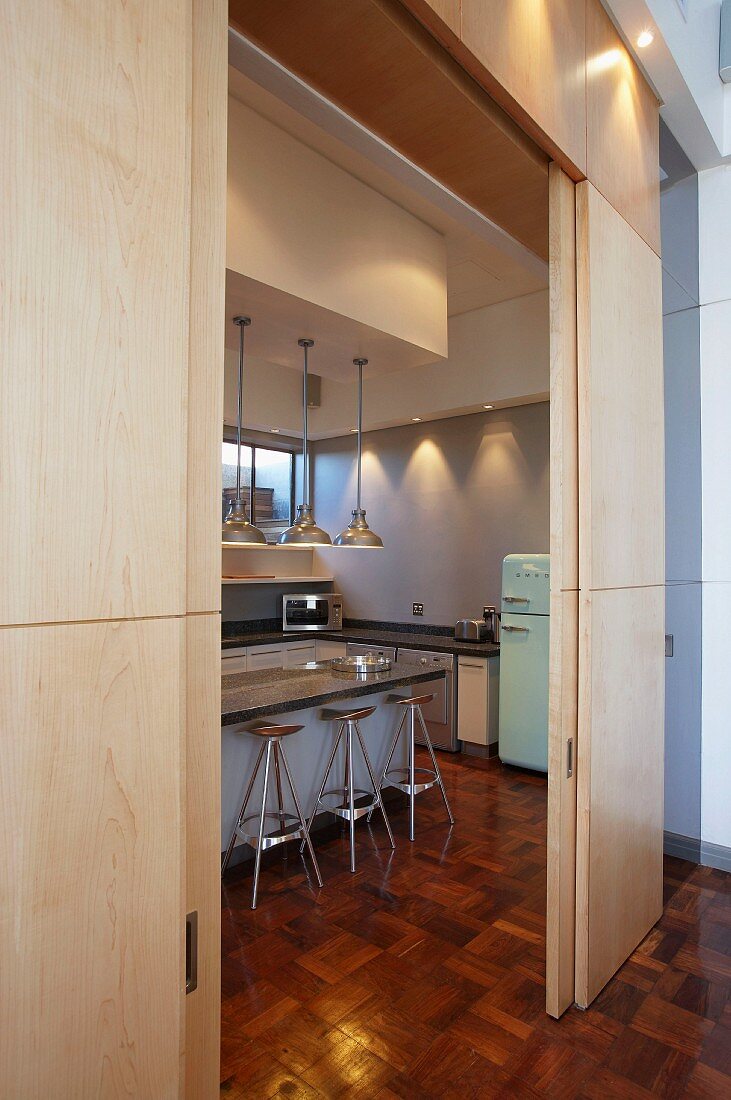 Blick durch offene Holz-Schiebeelemente auf Küchentheke mit Hängeleuchten, Barhockern und Kühlschrank