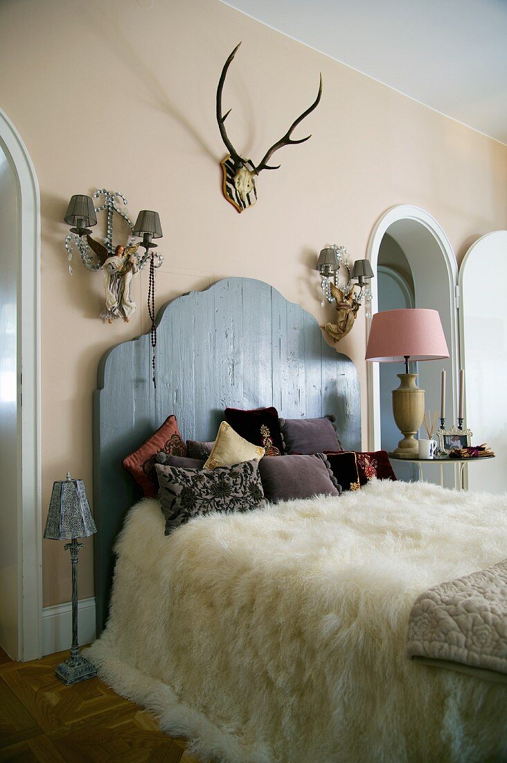 Weisses Tierfell auf französischem Bett mit rustikalem Holzkopfteil an Wand zwischen Wandleuchten und Geweih