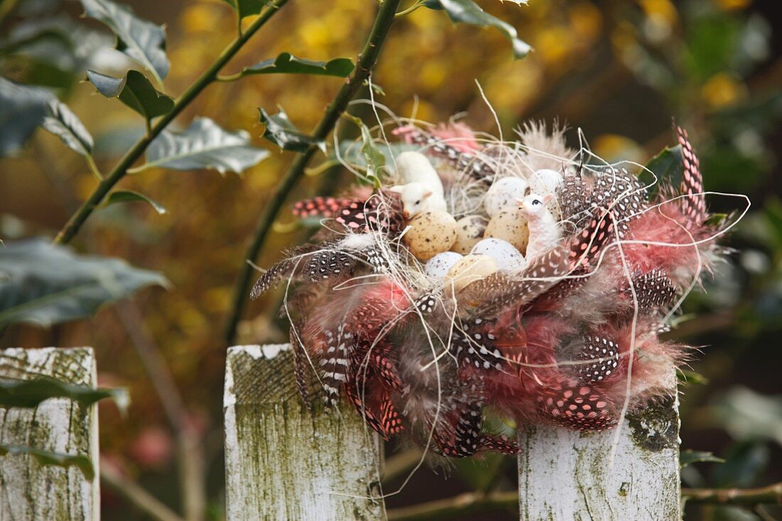 Zartes Osternest aus gepunkteten Federn mit Vogeleiern und Schäfchenfiguren auf einem Lattenzaun